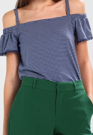Kvinde iført en blå og hvidstribet off-shoulder top med korte ærmer og høj taljede grønne bukser med lommer.