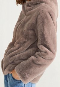 Femme portant une veste en fausse fourrure douce et légère de couleur marron clair avec des poches latérales et un jean bleu, vue de profil à hauteur de taille.