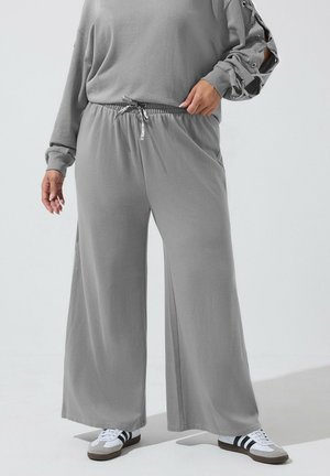 Personne portant un pantalon de survêtement gris ample avec une taille à cordon et un haut gris à manches lacées, associé à des baskets blanches avec des rayures noires.