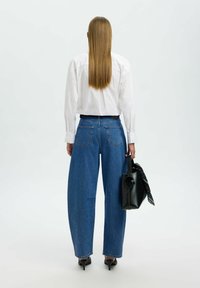 Camisa blanca abotonada, vaqueros azules de pierna ancha con bolsillos traseros, bolso negro con pañuelo estampado, tacones elegantes, cabello liso y largo.