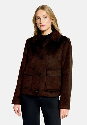 Femme portant une veste marron foncé boutonnée avec de grandes poches avant, un pantalon noir et un haut noir sur un fond clair uni.