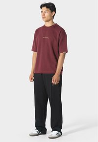 Bordová košile s krátkým rukávem a vyšitým textem na hrudi, spárovaná s černými kalhotami s zlatým textem a bílými teniskami.