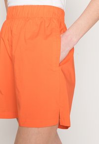 Shorts orange avec une taille élastique, des poches sur les côtés et une coupe droite. Le tissu semble lisse avec un léger éclat.