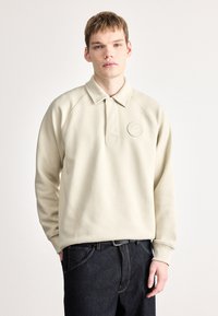 Krém színű, puha textúrájú galléros pulóver, gombolható nyakkal és hímzett címerrel, sötét farmernadrággal párosítva.