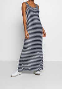 Frau in einem marineblauen und weißen gestreiften ärmellosen Maxikleid mit Knöpfen, kombiniert mit weißen Sneakers, vor einem einfarbigen Hintergrund stehend.