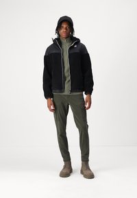 Schwarz-graue Fleecejacke mit Reißverschluss, grüne Mittelschicht, olivfarbene Cargo-Hosen und sandfarbene Stiefel. Robuste Verarbeitung mit verstellbaren Handgelenk-Bündchen.