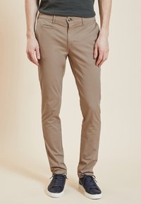 Chino beige en coton avec une coupe slim, poches avant et fermeture à bouton, associés à des baskets bleu foncé. Texture lisse.