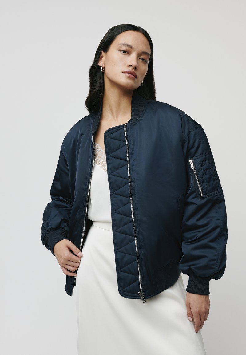 Giubbotto bomber navy con design trapuntato, polsini a coste e tasca con zip sulla manica. Realizzato in tessuto liscio e lucido.