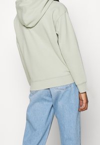 Jasnozielona bluza z kapturem o miękkiej fakturze, z kapturem sznurowanym i ściągaczem na dole. W zestawie z jasnoniebieskimi dżinsami.