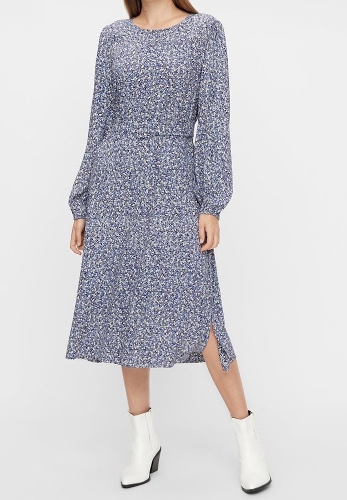 Femme portant une robe midi à manches longues bleue à motif floral avec une fente latérale, associée à des bottines blanches, sur un fond clair uni.