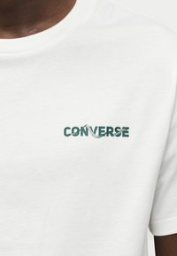 Λευκό βαμβακερό t-shirt με πράσινο γραφικό λογότυπο "CONVERSE" στο στήθος. Κοντά μανίκια με κλασικό στρογγυλό γιακά.