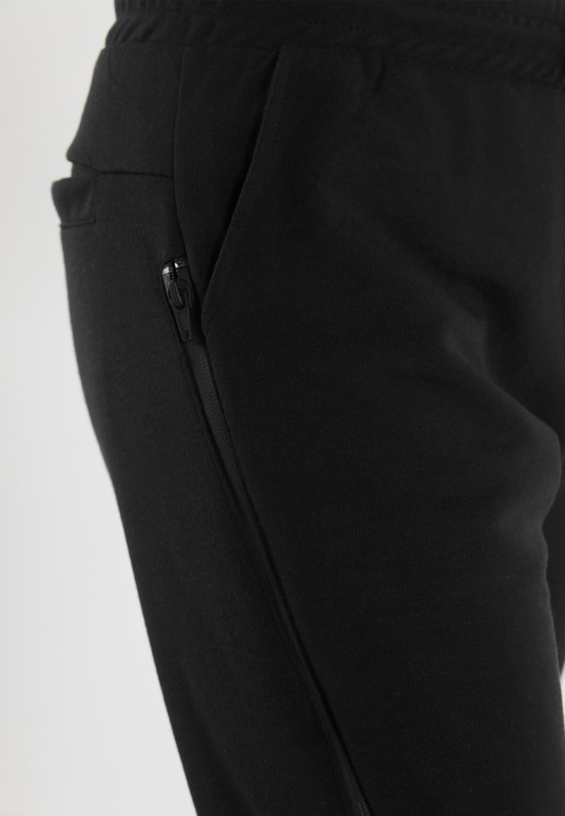 Mustat lenkkihousut, jotka on valmistettu pehmeästä kankaasta, sisältävät sivussa vetoketjullisen taskun ja joustavan vyötärön mukavuutta varten.