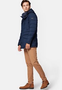 Granatowa ocieplana kurtka parka z zamkiem błyskawicznym i zapięciem na guziki, wieloma przednimi kieszeniami oraz odpinanym kapturem, noszona z jasnobrązowymi spodniami i butami.