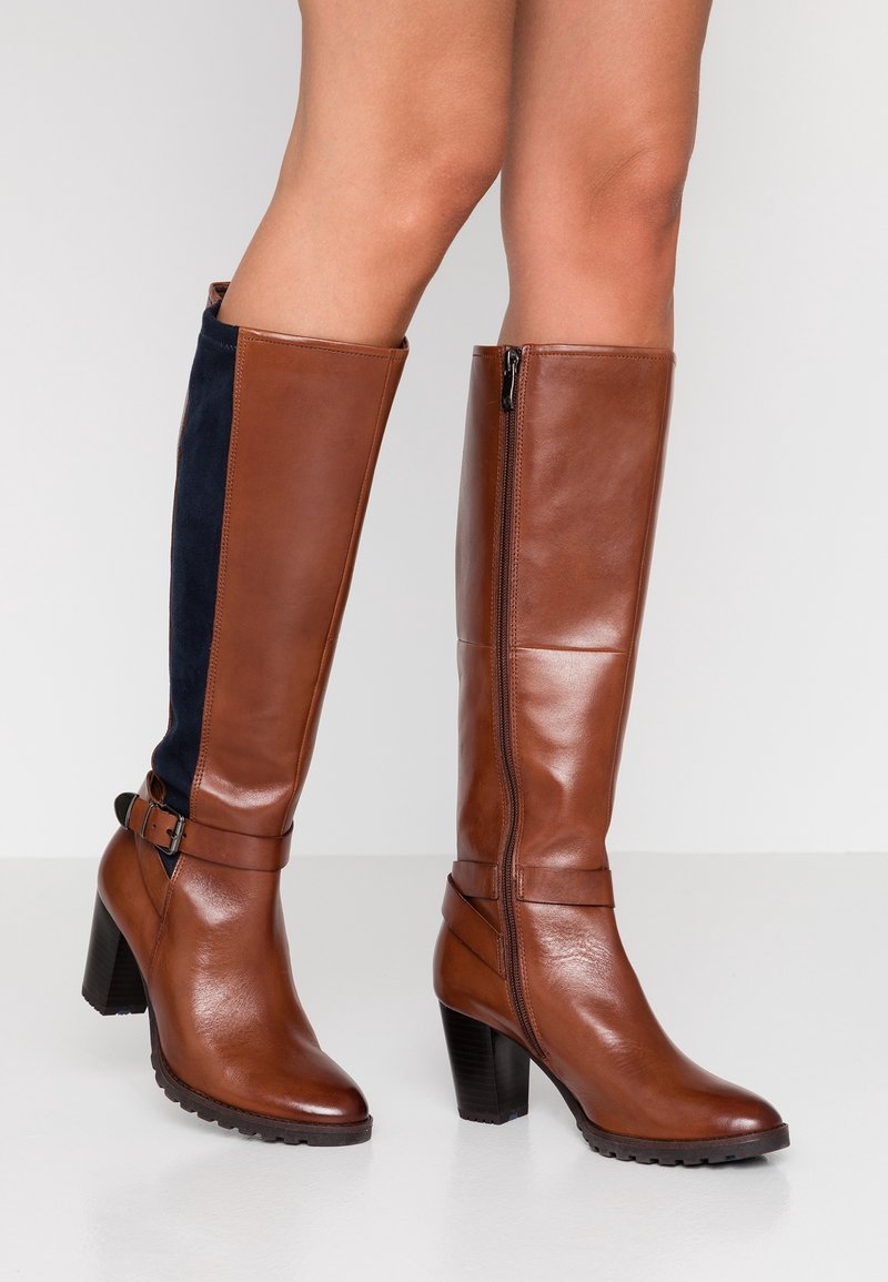 Botas altas marrones de cuero hasta la rodilla con un panel de ante azul marino, tacón alto, detalle de correa en el tobillo y cierre de cremallera lateral.