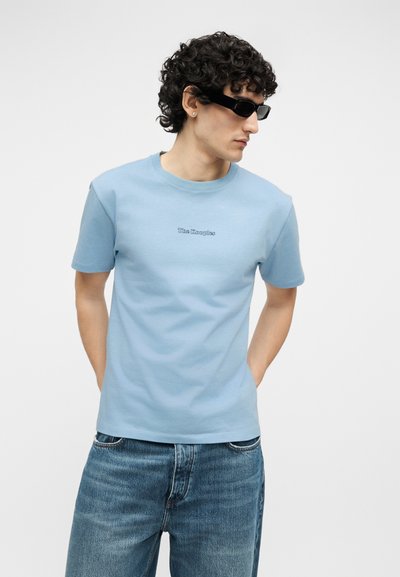 Άνδρας με σγουρά μαλλιά, που φοράει μαύρα γυαλιά ηλίου, ανοιχτό μπλε κοντομάνικο με το κείμενο "The Kooples" και μπλε τζιν, στέκεται με τα χέρια πίσω από την πλάτη.