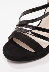 Sandale noire à bout ouvert avec plusieurs lanières brillantes à texture crocodile et une lanière en daim sur les orteils sur un fond clair.
