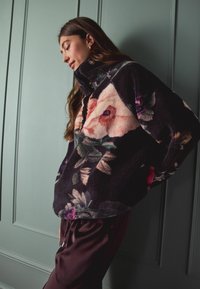 Kwiatowy polarowy pulower w ciemnych kolorach, z wysokim kołnierzem i miękką fakturą. Podkreślony różowymi i zielonymi kwiatowymi wzorami.