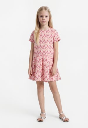 Młoda dziewczyna stojąca, ubrana w krótki różowy sukienkę w wzór jodełki i białe sandały na jednolitym białym tle.