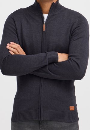 Homme portant un pull zippé gris foncé avec un col et des poignets côtelés, les bras croisés, avec un patch logo marron visible près de l'ourlet.