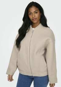 Bež zip-up bomber jakk, millel on tekstureeritud kangas, laiad varrukad ja ümar krae, koos siniste teksadega.