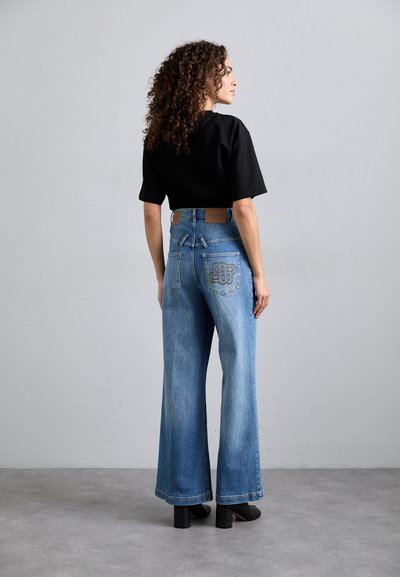 Széles lábú, világoskék farmernadrág magas derékkal, barna bőr címkével és díszvarrással a hátsó zsebeken. Fekete inggel viselve.