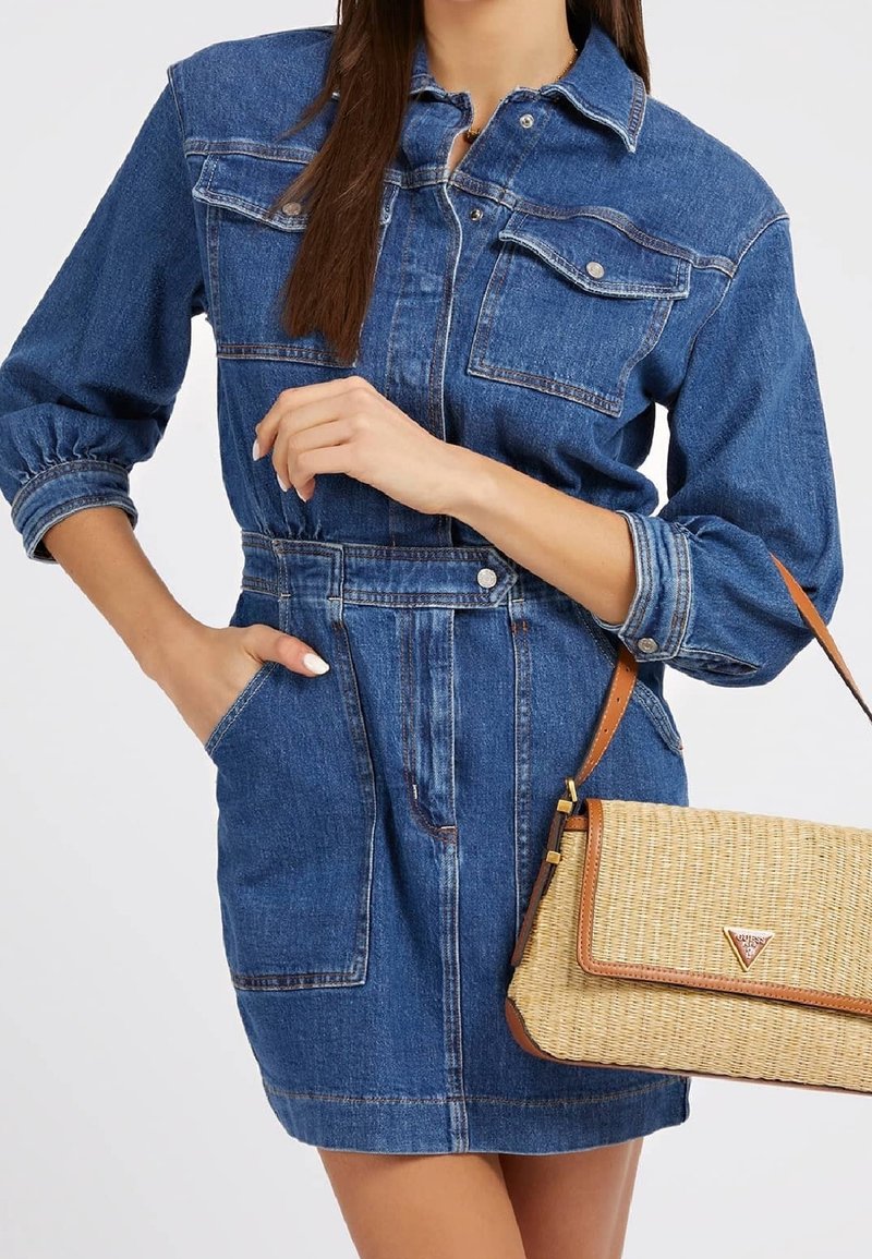 Femme portant une robe en denim bleu avec manches retroussées, tenant un sac à main tissé beige avec des garnitures en cuir marron.