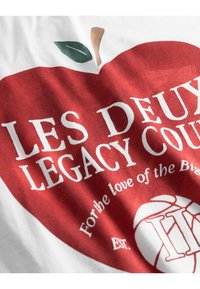 Červený grafický design s stylizovaným jablkem a zeleným listem. Text zahrnuje "LES DEUX LEGACY COURT" a "Pro lásku k velkým věcem". Bavlněný materiál.
