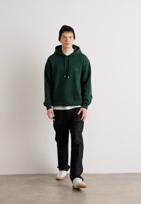 Sudadera de forro polar verde con bolsillo canguro y cordón, combinada con jeans negros y zapatillas blancas. Usada con un gorro negro.
