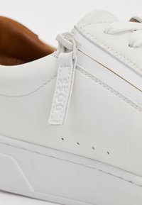 Baltas ādas sneakers ar gludu tekstūru, kas aprīkots ar sānisko rāvējslēdzēju, izšūtiem akcentiem un minimālām perforācijām ap zoli.