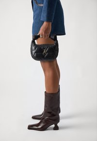 Black leather handbag with quilted design and gold insect emblem, held by a person wearing a blue coat and brown knee-high boots.