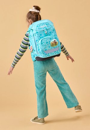 Child wearing large turquoise backpack with heart patterns and horse patches, balancing on one foot against beige background.