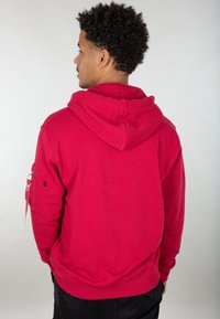 Felpa rossa con cappuccio e finitura testurizzata, dotata di tasca laterale e dettagli con zip. Progettata con maniche lunghe e una vestibilità rilassata.