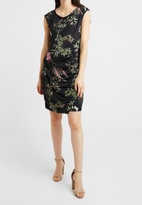 Kobieta nosząca bezrękawową czarną sukienkę w zielony i różowy kwiatowy wzór, z marszczeniem po boku, oraz beżowe sandały na obcasie z paskiem wokół kostki.