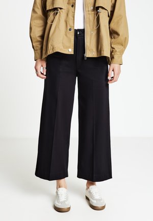 Pantalon noir à jambes larges, à la texture lisse, avec une taille haute et des plis. Associé à une veste kaki zippée et des baskets blanches.