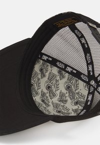 Svart mesh truckerkeps med en böjd skärm. Inuti finns en grå foder med svarta ögon- och vinggrafikmönster. Von Dutch-logotyp synlig.