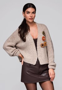 Beige pletený cardigan s dlouhými rukávy, který má tři vyšívané květinové akcenty v oranžové, zelené a hnědé barvě. V kombinaci s hnědou koženou sukní.