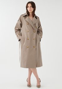 Gabardina beige de doble botonadura, fabricada en una mezcla de algodón, con botones en tono dorado, grandes bolsillos y cinturón en la cintura. Textura suave.