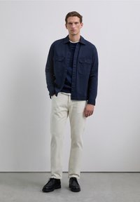 Camisa de botones azul marino con dos bolsillos en el pecho, sobre un suéter azul marino, combinada con pantalones crema y zapatos negros. Textura de tela suave.