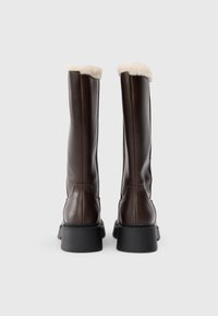 Bottes en cuir marron jusqu'au genou avec une douce doublure en fourrure crème au sommet et une semelle épaisse noire. Présente un design épuré et un minimum de coutures.