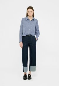 Camisa de botones a rayas azul y blanca combinada con pantalones de mezclilla oscura de pierna ancha con un puño de mezclilla clara. Mocasines negros completan el atuendo.