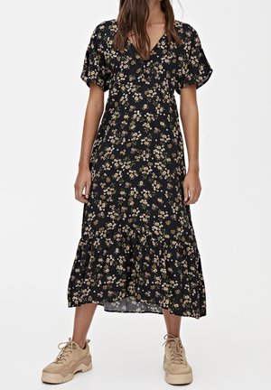 Femme portant une robe midi noire avec de petits imprimés floraux blancs et jaunes, et des baskets beige à semelle épaisse, debout devant un fond blanc.