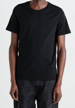 T-shirt en coton noir avec un col rond et des manches courtes. Design simple, texture lisse, porté avec un pantalon sombre à côtes.