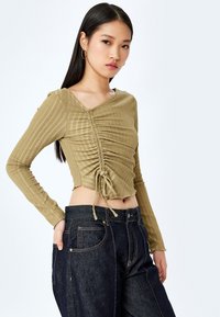 Olijfgroene cropped longsleeve top van textiel met een trekkoorddetail op de taille, gecombineerd met donkerblauwe high-waisted jeans.