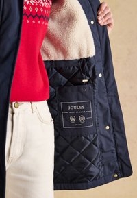 Persona con maglione rosso e pantaloni bianchi tiene aperta una giacca trapuntata blu navy foderata in pile crema, mostrando l'etichetta del marchio Joules all'interno.