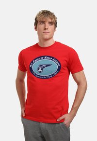 Rød bomulds t-shirt med et cirkulært grafik, der viser "Pan American World Airways," et blåt flydesign og tekst nedenunder. Kortærmet, rund hals.