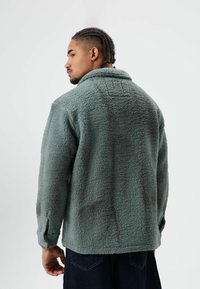 Groene fleecejas met een gestructureerde, pluizige afwerking, knoopsluiting aan de voorkant en een kraag, met een embleem op de achterkant.