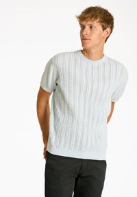 Ljusblå stickad tröja med en texturerad vågmönster, korta ärmar och rundad halsringning, kombinerad med svarta byxor.