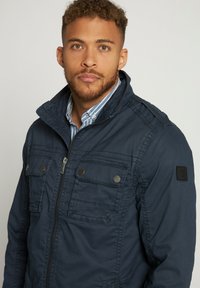 Donkerblauwe jas met een ritssluiting aan de voorkant, twee borstzakken en een zwart schouderpatch. Voorzien van een gestructureerde kraag en een contrasterende blauwgestreepte shirt eronder.