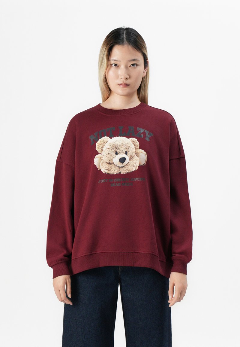 Young person wearing burgundy sweatshirt with teddy bear graphic and text "NOT LAZY" and dark blue pants, standing against white background.