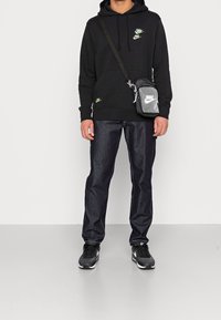 Svart huvtröja med grön Nike-logga, mörka jeans och svarta sneakers. Inkluderar en grå axelväska med reflekterande detaljer.